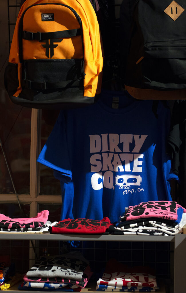 Photo of a shop display with folded t-shirts and a yellow backpack hanging up. One shirt is hanging prominently behind the shelf. It is a vibrant blue and says "dirty skate co." in white lettering with a cartoon drawing of a skull, also in white.