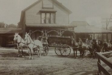 old fashioned photo of a team of white horses pulling a hearse down aa wide dirt street with a single building in the background.