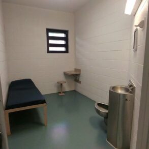 Photo of the inside of a jail cell, with a bed, sink and toilet in a small white room