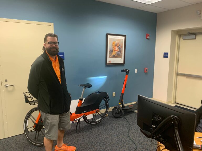 Image of a man indoors next to an electric bicycle and an electric scooter