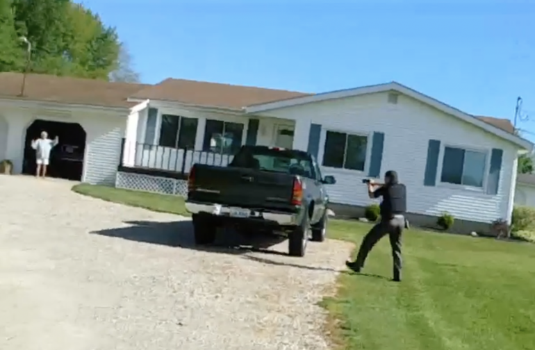 Image of a deputy moving toward a woman with his gun aimed at her while she is standing in the entryway of a garage