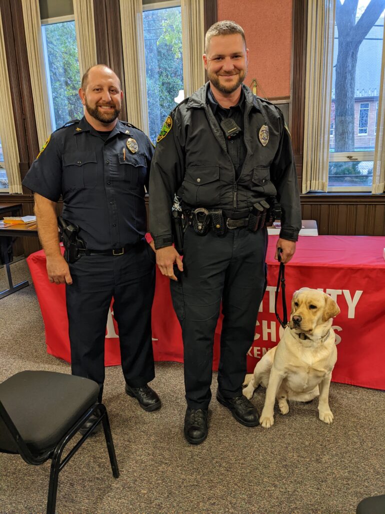 Two police officers and a yellow lab on a leash pose for a picture indoors