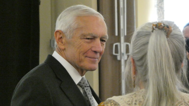 Image of an older white man with gray hair in a suit smiling and engaging with attendees of his event