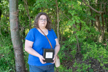 A woman in a blue shirt stands in the woods holding a black box with a label on it